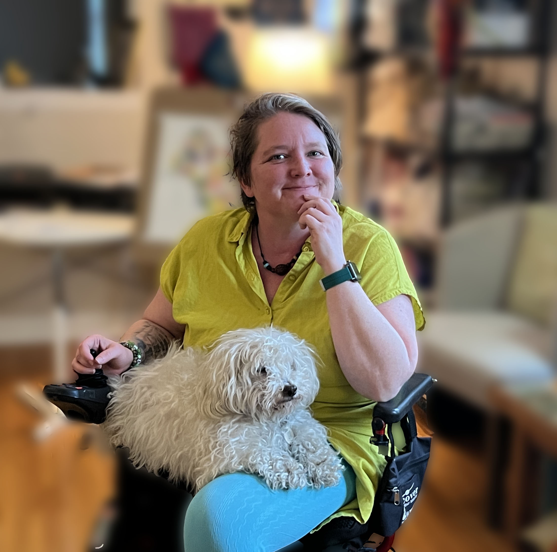 Mallory sitting in her art studio with Dodger, a small white dog, in her lap- soft indoor lighting behind her with a window to the right just out of view.  The background is blurred but Mallory and Dodger are in focus.
