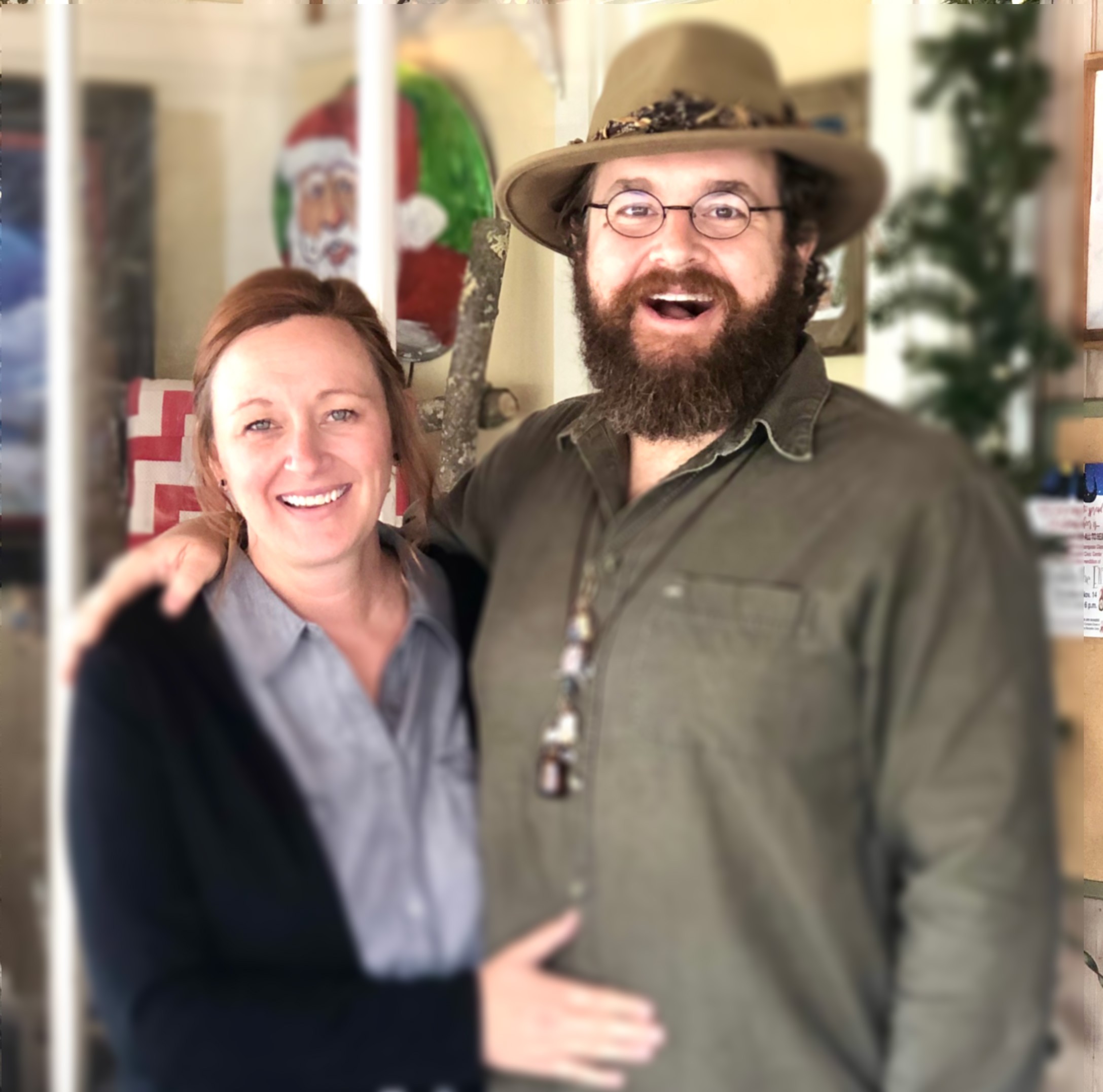 Mallory sitting in her art studio with Dodger, a small white dog, in her lap- soft indoor lighting behind her with a window to the right just out of view.  The background is blurred but Mallory and Dodger are in focus. 